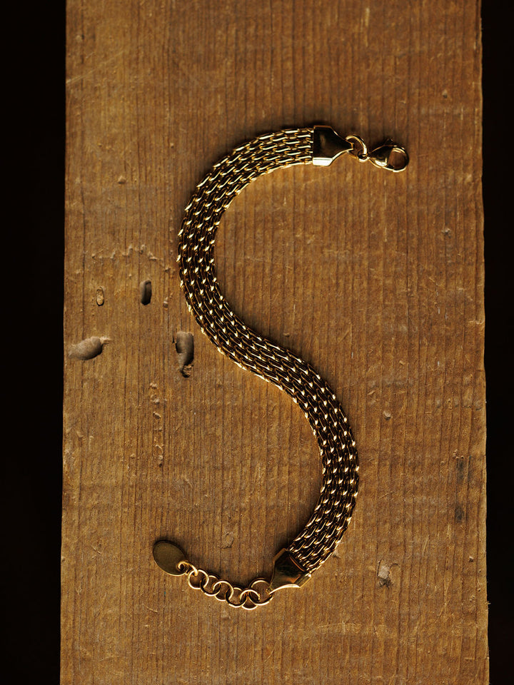 The Vienna Bracelet by Savannah Sevens western life{&}style features multiple strands of 18K gold-filled metal braided in an S-shape, displayed on a rustic wooden surface.