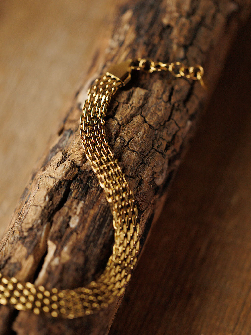 The Vienna Bracelet by Savannah Sevens western life{&}style—a hypoallergenic, woven gold braided design—rests on textured wood, showcasing its intricate links and polished 18K gold-filled finish.