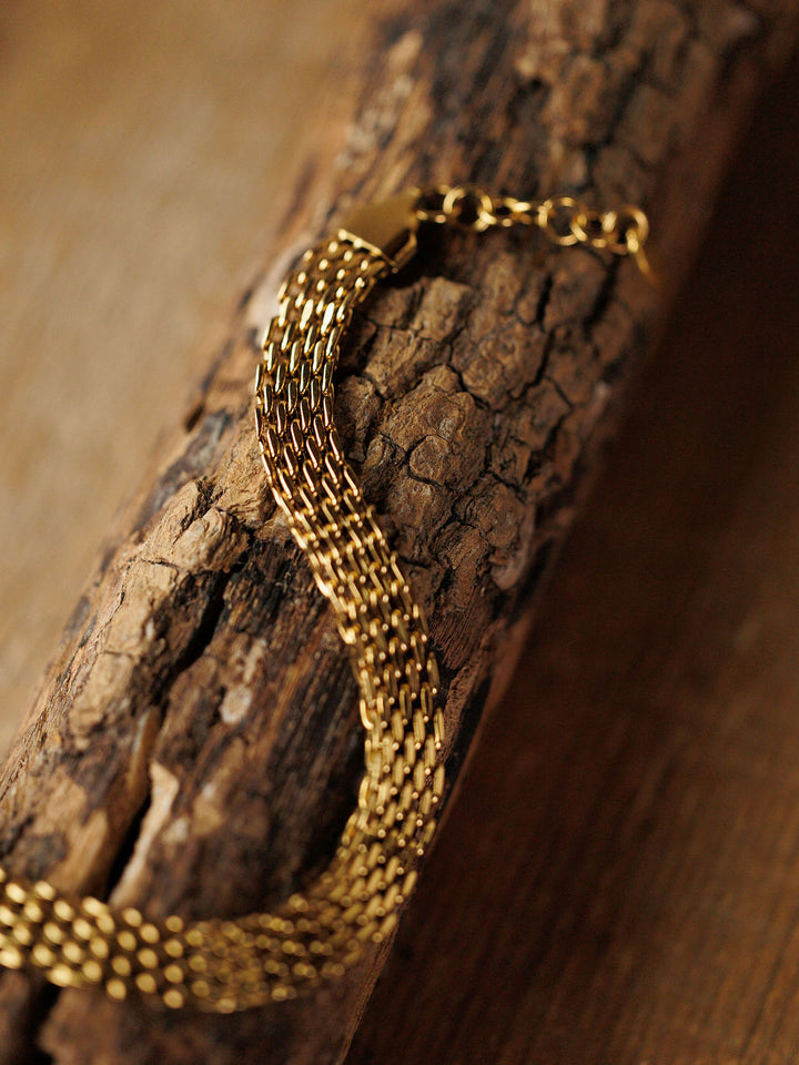 The Vienna Bracelet by Savannah Sevens western life{&}style—a hypoallergenic, woven gold braided design—rests on textured wood, showcasing its intricate links and polished 18K gold-filled finish.