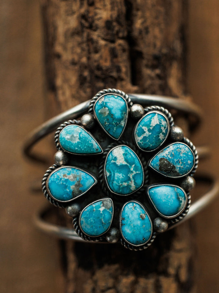 The Sagola Cuff by Savannah Sevens western life{&}style is a unique sterling silver cuff with a floral design, showcasing eight blue turquoise stones encircling a central turquoise stone, set against a rustic wooden background.
