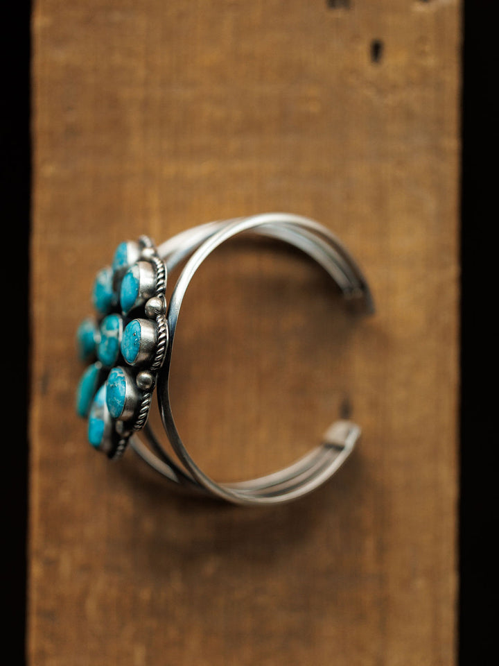 The Sagola Cuff by Savannah Sevens western life{&}style is a unique sterling silver bracelet with a cluster of blue turquoise stones, elegantly showcased on a rustic wooden surface.