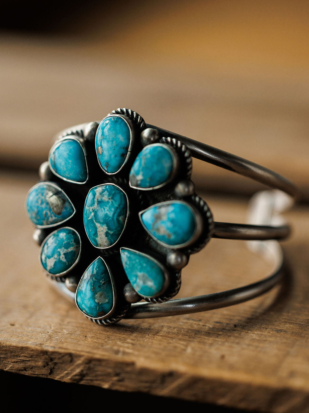 The Sagola Cuff by Savannah Sevens western life{&}style is a unique sterling silver bracelet with a central floral design and oval and teardrop turquoise stones, shown on a wooden surface.