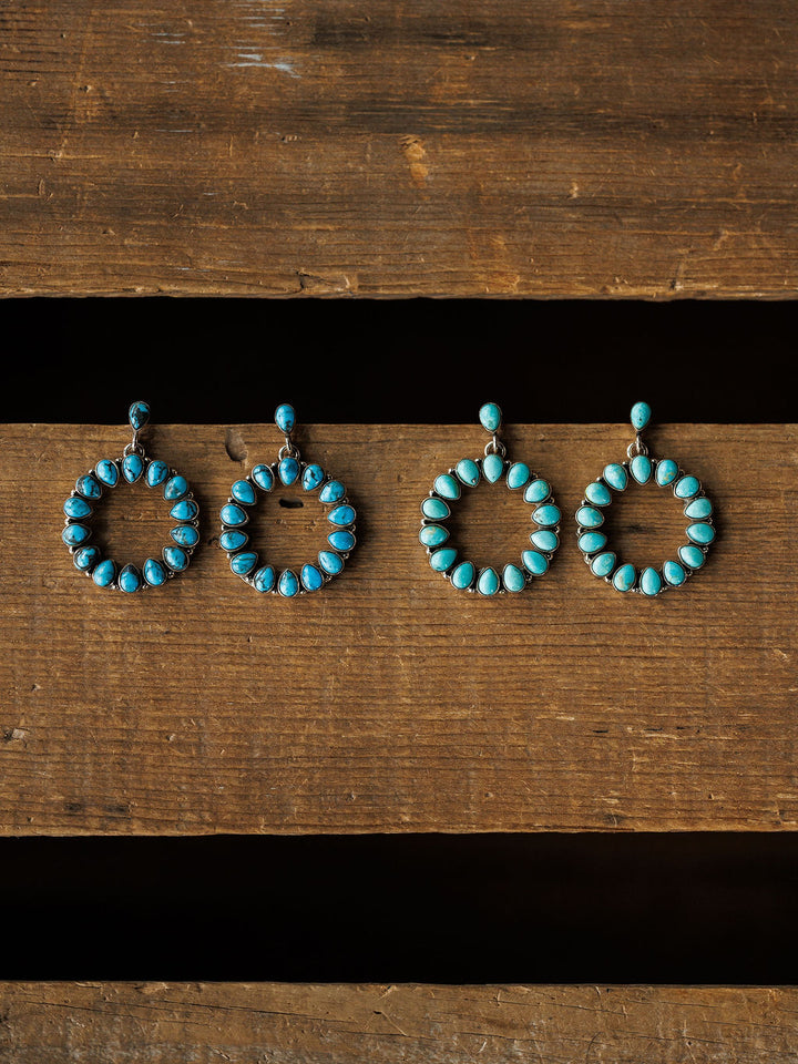 Four Ocala Earrings by Savannah Sevens western life{&}style are arranged in a row on rustic wood. Each sterling silver drop earring showcases blue and green Kingman turquoise with a single stone connecting the hoop to the post.