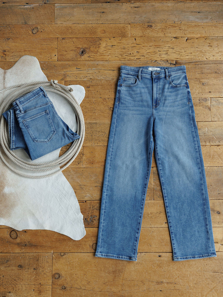 A pair of blue jeans lies flat on a wooden floor beside a folded Savannah Sevens western life{&}style The Layton High Rise Relaxed Straight Leg Jean, a coiled rope, and a cowhide rug.