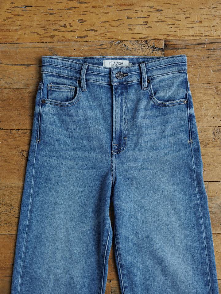 The Layton High Rise Relaxed Straight Leg Jean by Savannah Sevens western life{&}style, in light blue wash with button and zipper fly, front and coin pockets, and brand label on the waistband, laid flat on a wooden surface.