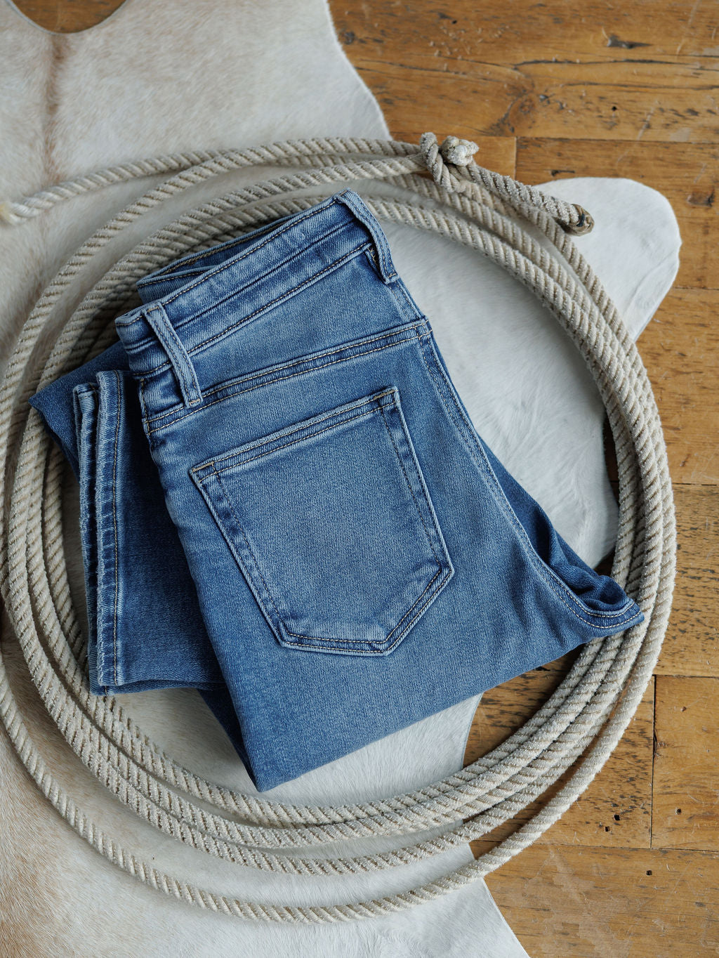 The Layton High Rise Relaxed Straight Leg Jean by Savannah Sevens western life{&}style, folded in blue, rests inside a coiled rope atop a white animal hide on a wooden floor.