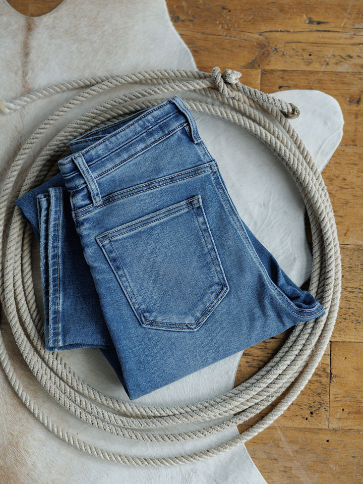 The Layton High Rise Relaxed Straight Leg Jean by Savannah Sevens western life{&}style, folded in blue, rests inside a coiled rope atop a white animal hide on a wooden floor.