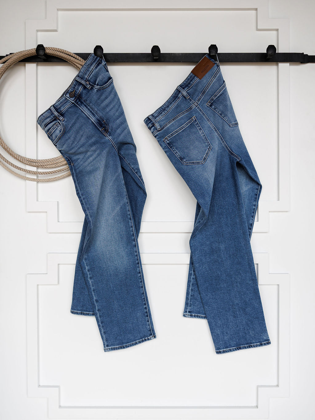 Two pairs of The Layton High Rise Relaxed Straight Leg Jeans by Savannah Sevens western life{&}style hang side by side on a black rod against a white wall with decorative molding; a coiled rope hangs on the left.
