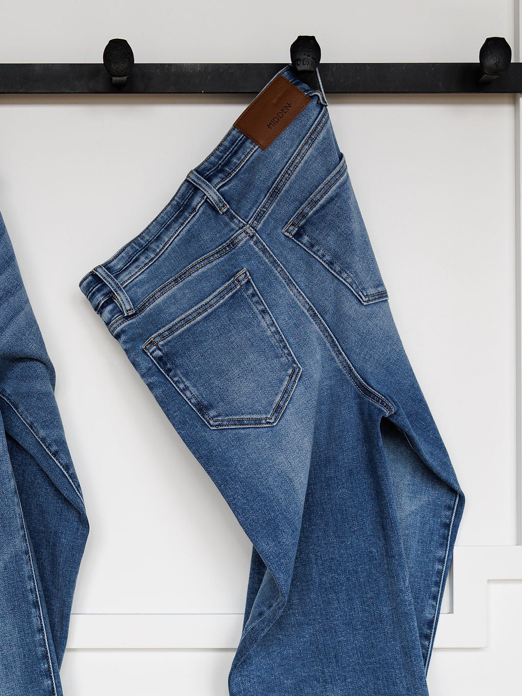The Layton High Rise Relaxed Straight Leg Jean by Savannah Sevens western life{&}style, featuring visible back pockets and a brown leather brand patch, hangs on a black wall hook against a white background.