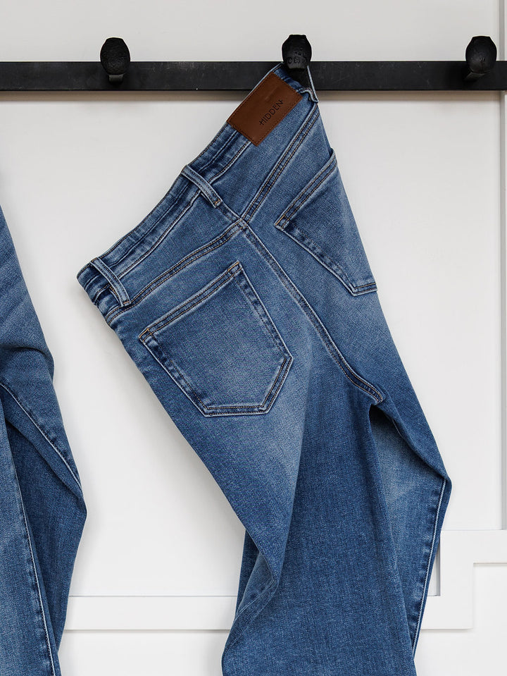 The Layton High Rise Relaxed Straight Leg Jean by Savannah Sevens western life{&}style, featuring visible back pockets and a brown leather brand patch, hangs on a black wall hook against a white background.