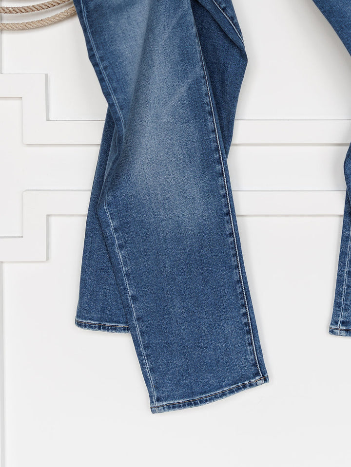 A close-up of the lower legs of The Layton High Rise Relaxed Straight Leg Jean by Savannah Sevens western life{&}style, hanging against a white wall with decorative paneling.