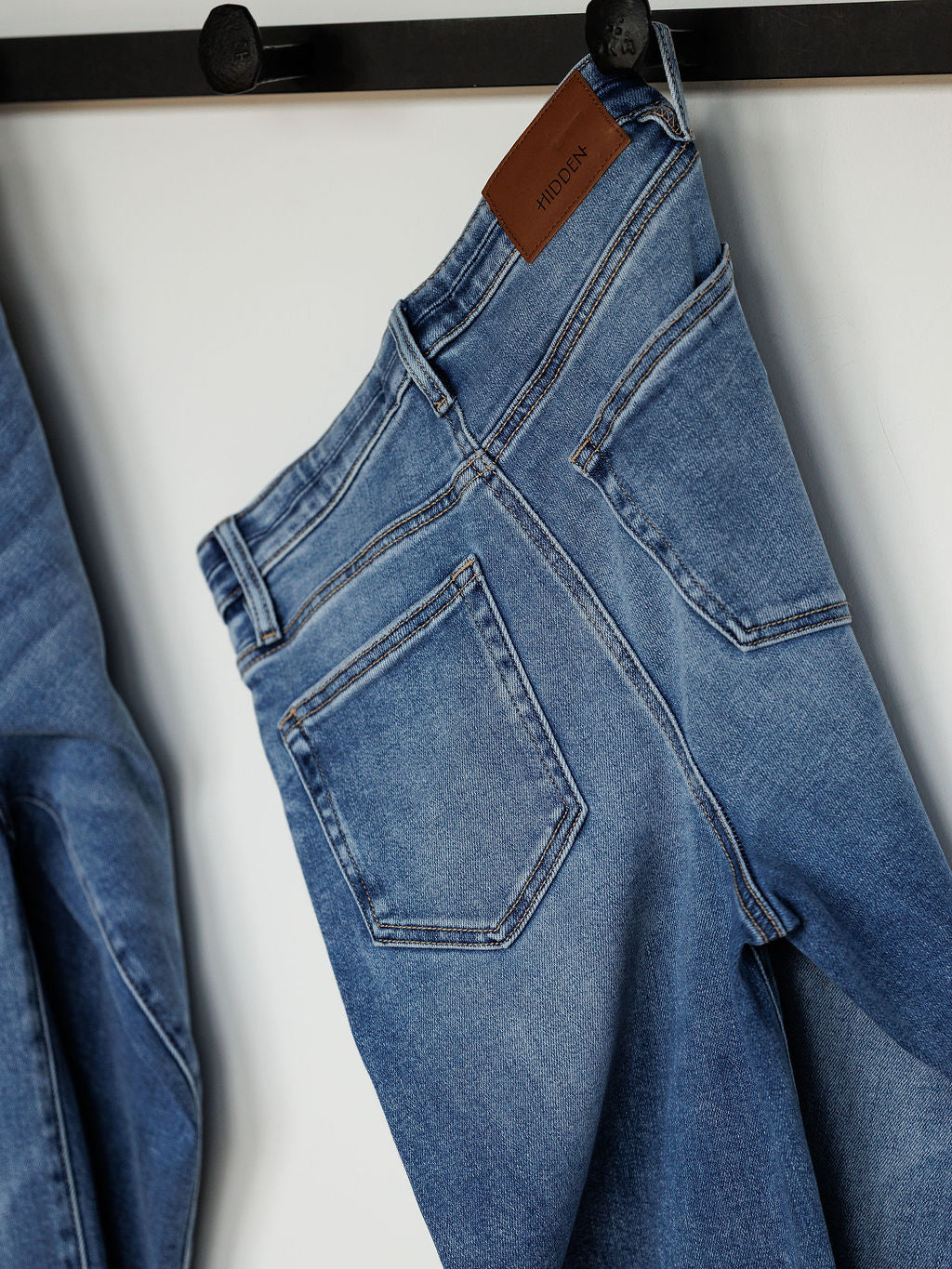 The Layton High Rise Relaxed Straight Leg Jean by Savannah Sevens western life{&}style, in blue denim, hangs by the waistband on a black rack, displaying the back pockets and a brown leather patch label.