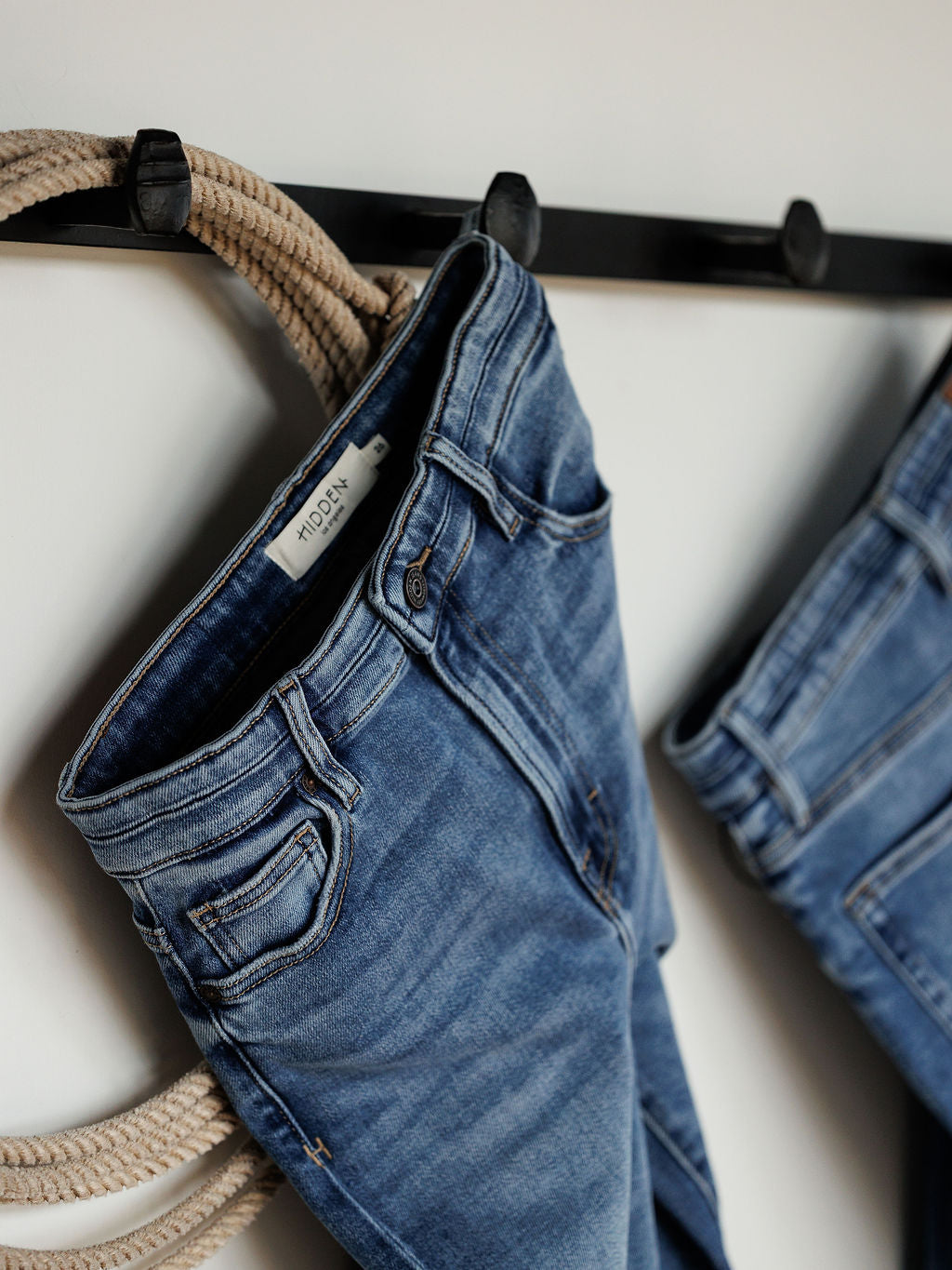 A pair of The Layton High Rise Relaxed Straight Leg Jeans by Savannah Sevens western life{&}style hangs on a black wall hook with a tan rope through the belt loops, while another relaxed straight leg jean is partially visible beside it.