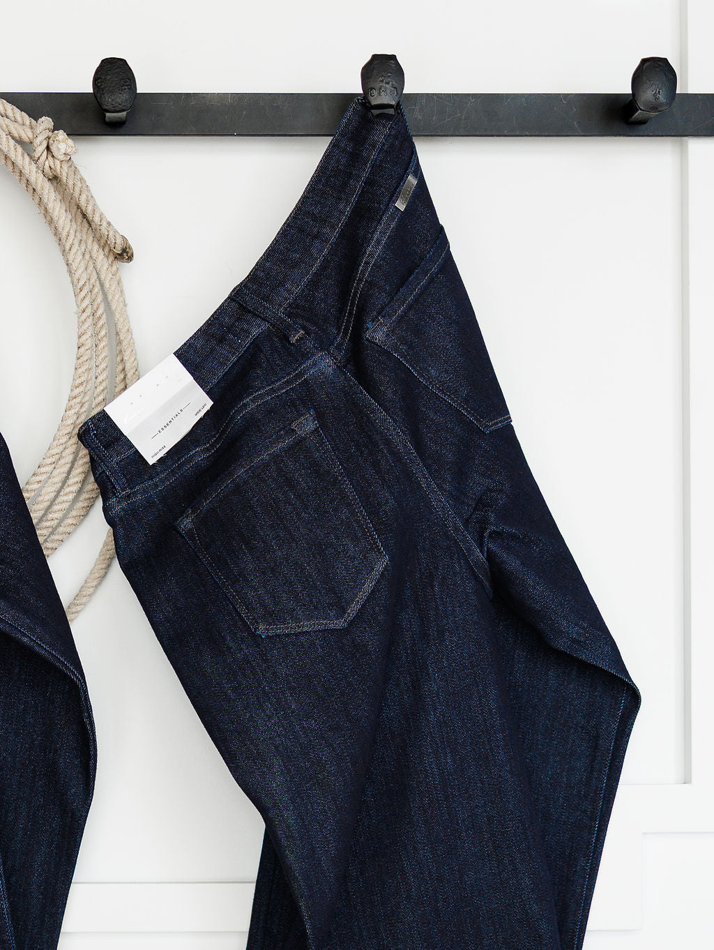 The Brody High Rise Wide Leg Jean by Savannah Sevens western life{&}style hangs on a black wall hook rack next to a coiled rope against a white wall.