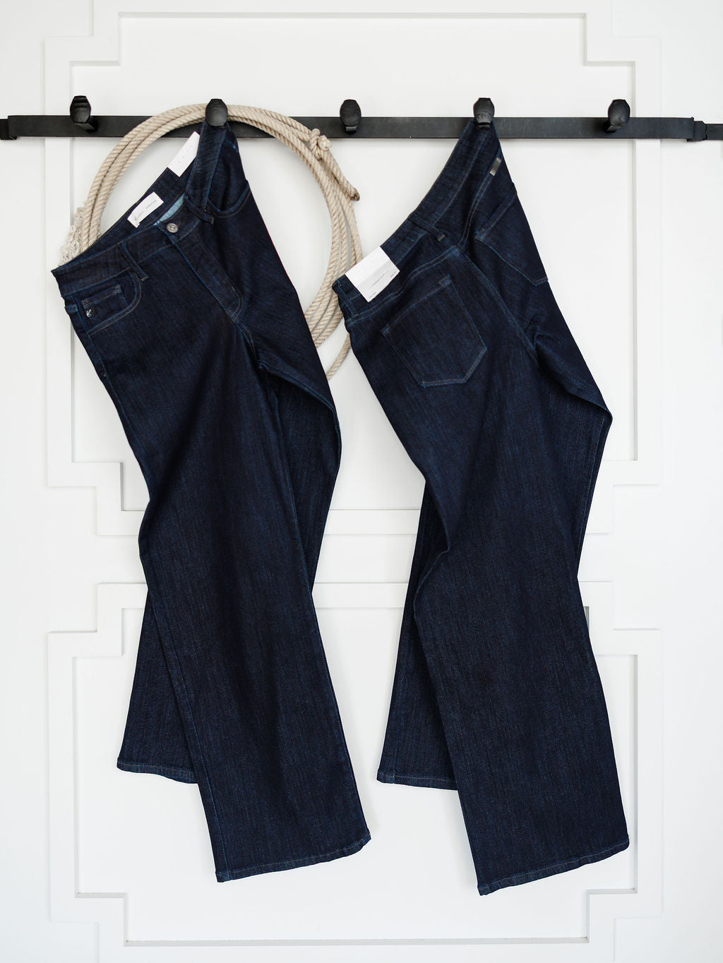 Two pairs of dark wash denim jeans, including The Brody High Rise Wide Leg Jean from Savannah Sevens western life{&}style, hang on a black wall-mounted rack with a coiled rope in the background against a white decorative wall.