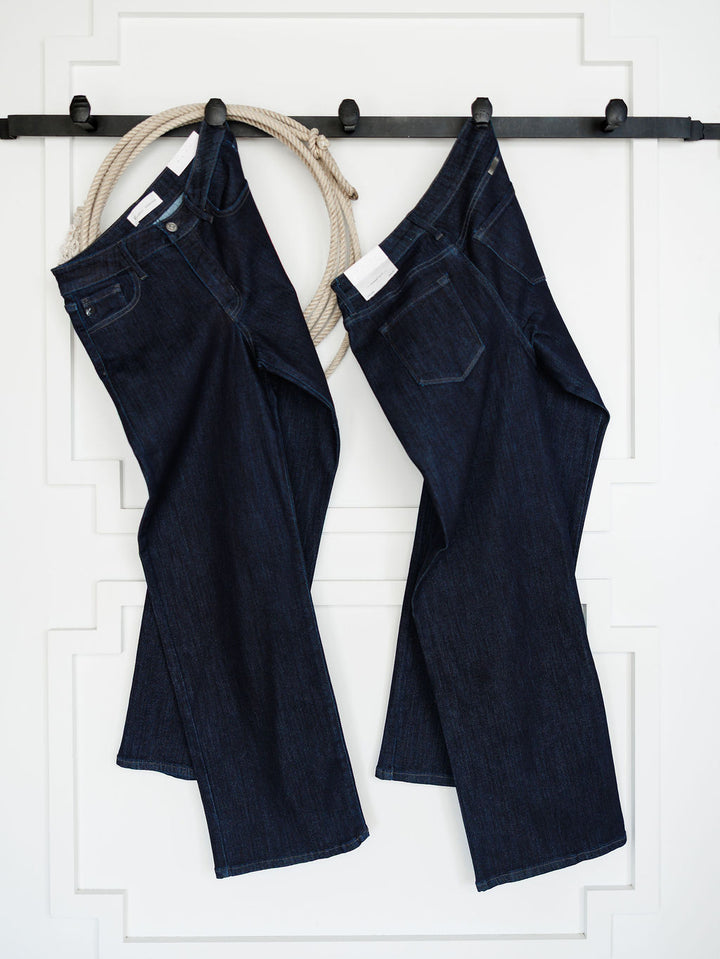 Two pairs of dark wash denim jeans, including The Brody High Rise Wide Leg Jean from Savannah Sevens western life{&}style, hang on a black wall-mounted rack with a coiled rope in the background against a white decorative wall.