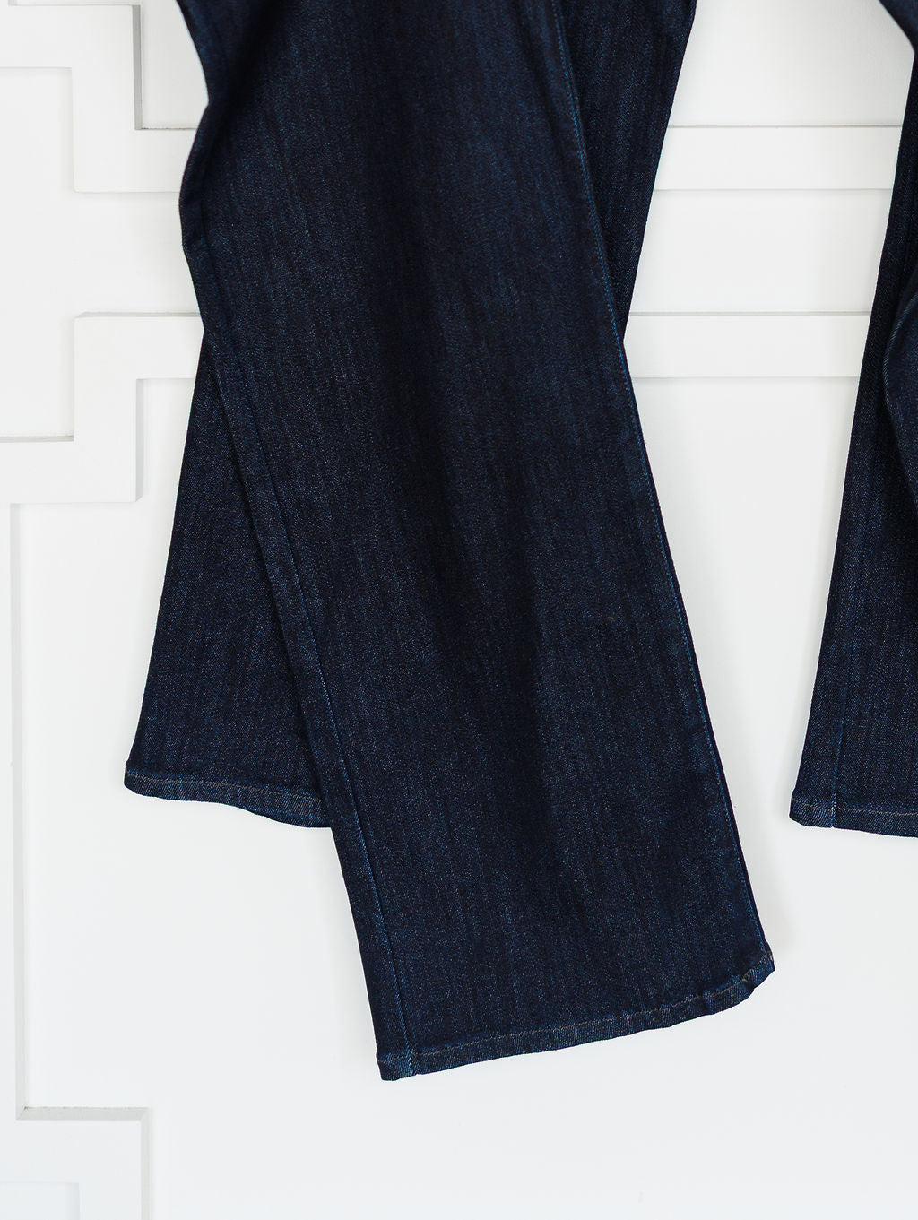 Close-up of The Brody High Rise Wide Leg Jean by Savannah Sevens western life{&}style, shown in dark wash denim with a slight flare, visible hem stitching, and wide leg cut against a white textured wall.