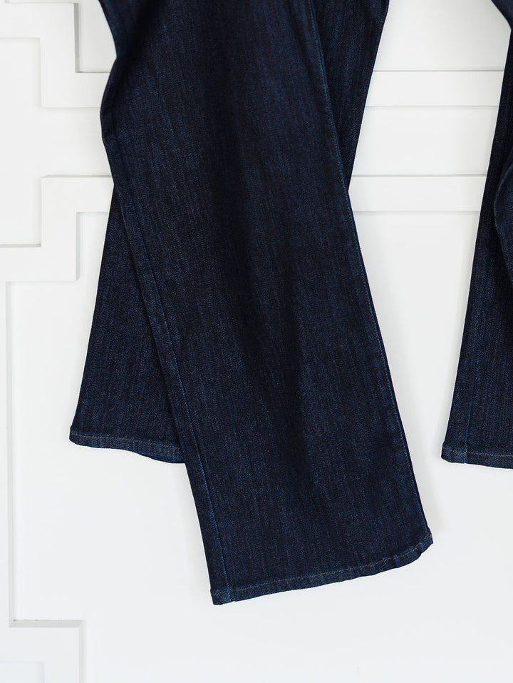 Close-up of The Brody High Rise Wide Leg Jean by Savannah Sevens western life{&}style, shown in dark wash denim with a slight flare, visible hem stitching, and wide leg cut against a white textured wall.