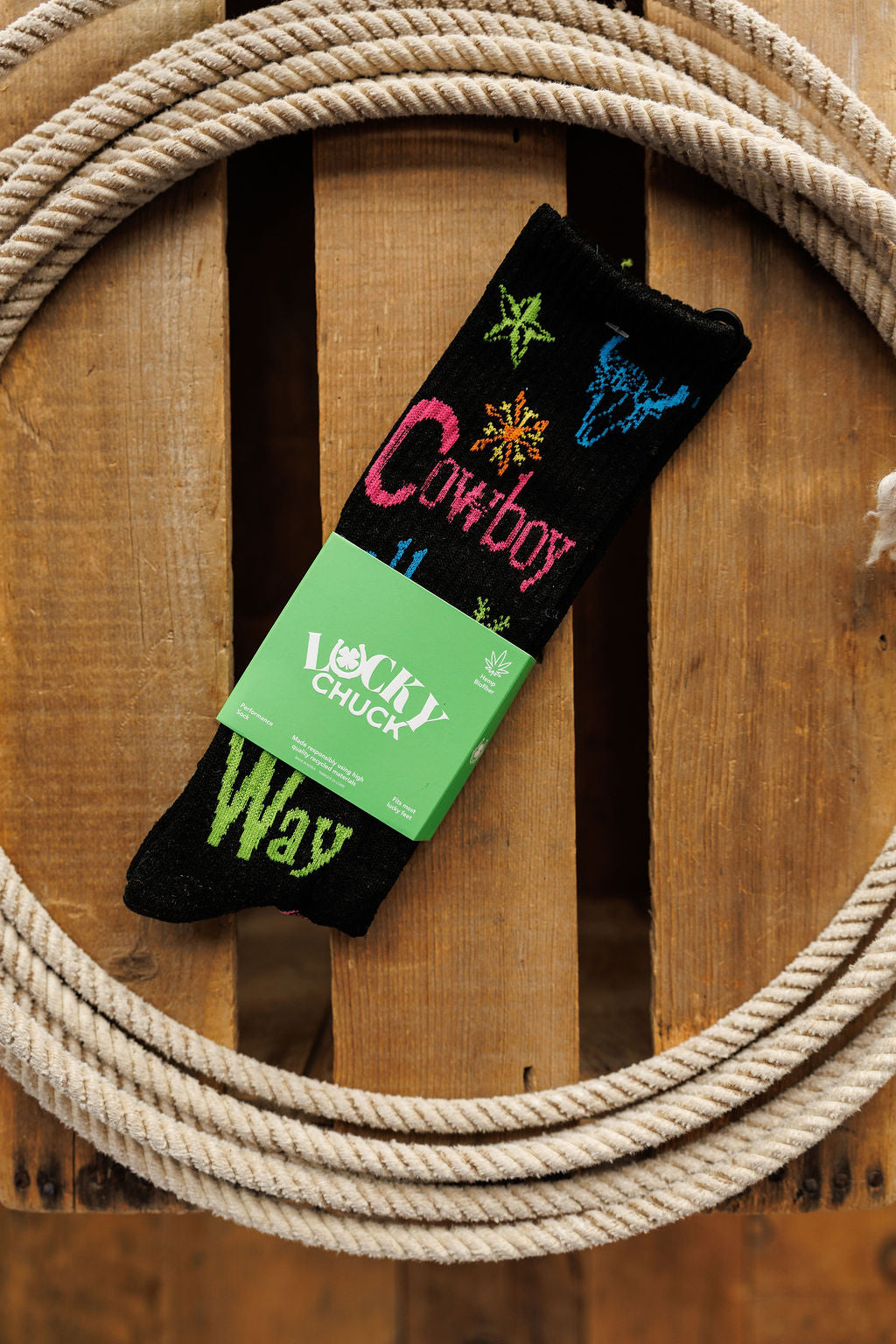 Cowboy All The Way Crew Socks by Savannah Sevens western life{&}style are black crew socks with colorful cowboy designs, made of an odor-fighting hemp blend, packaged with a green label on a wooden surface framed by coiled rope.
