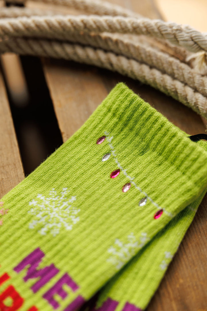 A close-up of Savannah Sevens western life{&}style’s Merry Christmas Y’all Crew Socks {Green} shows white snowflakes, red and silver bead accents, “MERRY” lettering, and a coiled rope on wood, highlighting this festive breathable sock.