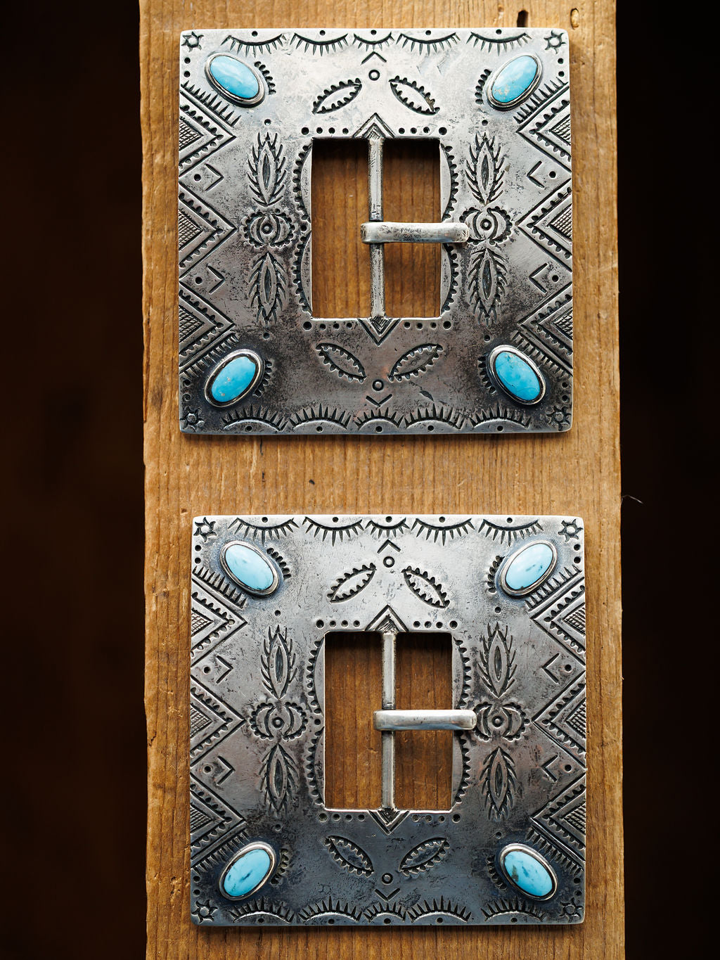 Two ornate, square Coalinga Buckles by Savannah Sevens western life{&}style, featuring engraved sterling silver and turquoise stones at each corner, are displayed vertically on a wooden plank.