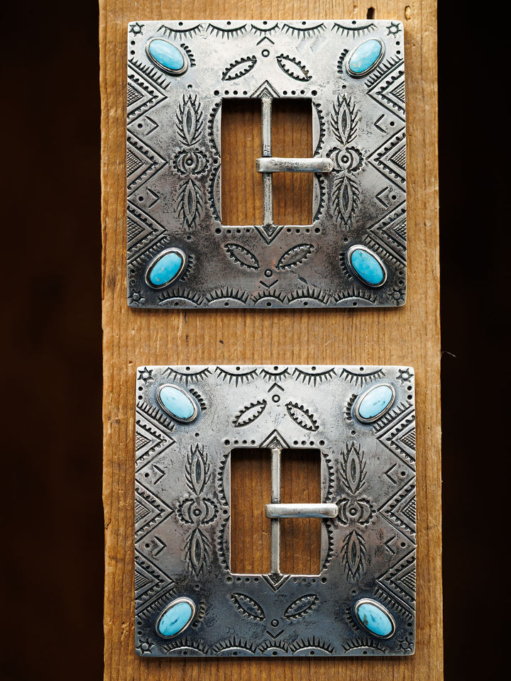 Two ornate, square Coalinga Buckles by Savannah Sevens western life{&}style, featuring engraved sterling silver and turquoise stones at each corner, are displayed vertically on a wooden plank.