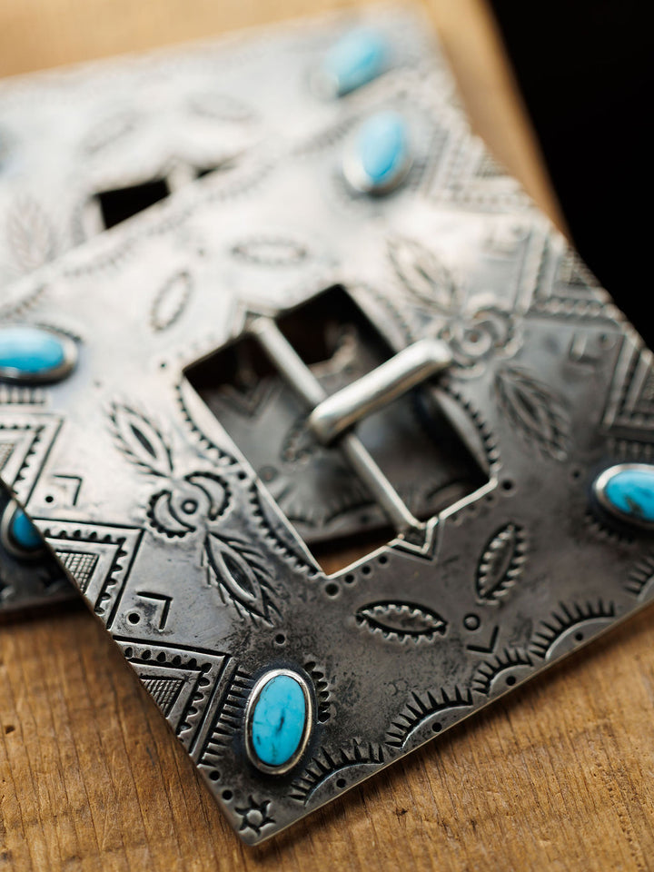 A close-up of the Coalinga Buckle by Savannah Sevens western life{&}style, a handmade sterling silver belt buckle with intricate engravings and oval turquoise stones at the corners, displayed on a wooden surface.
