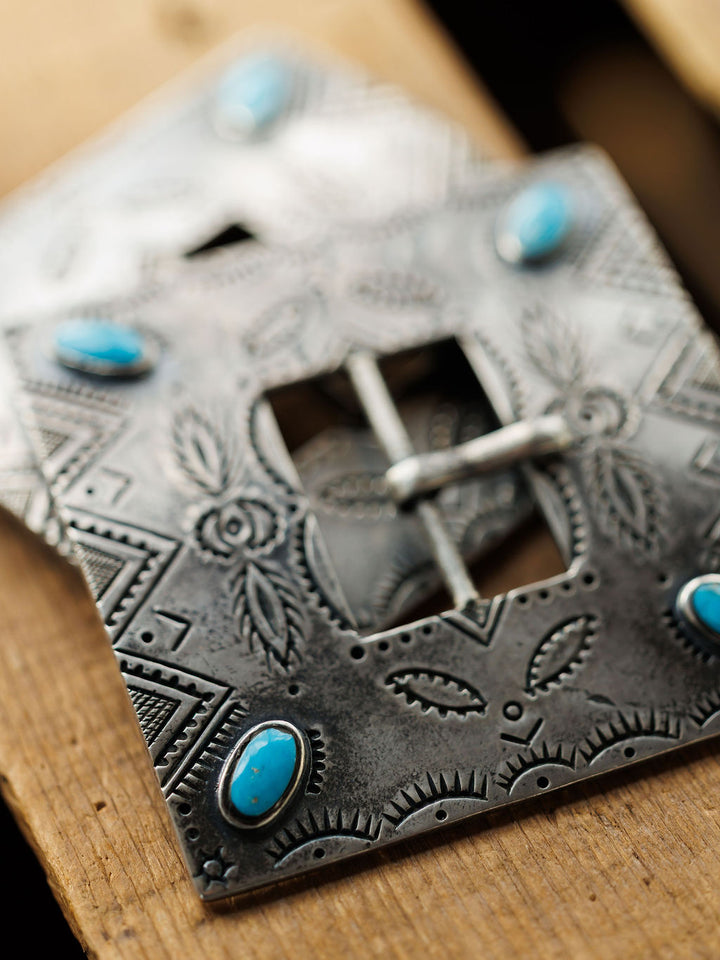 A close-up of the Coalinga Buckle by Savannah Sevens western life{&}style, a handmade sterling silver belt buckle with engraved patterns and four oval turquoise stones, shown resting on a wooden surface.