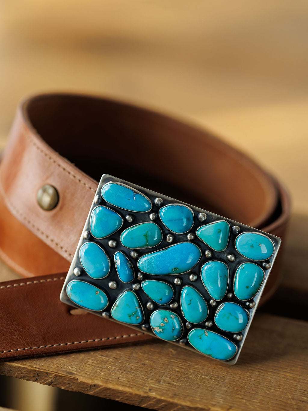 The Amorita Buckle by Savannah Sevens western life{&}style: a brown leather belt with a sterling silver buckle set with irregular Sonoran turquoise stones, displayed on a wooden surface.