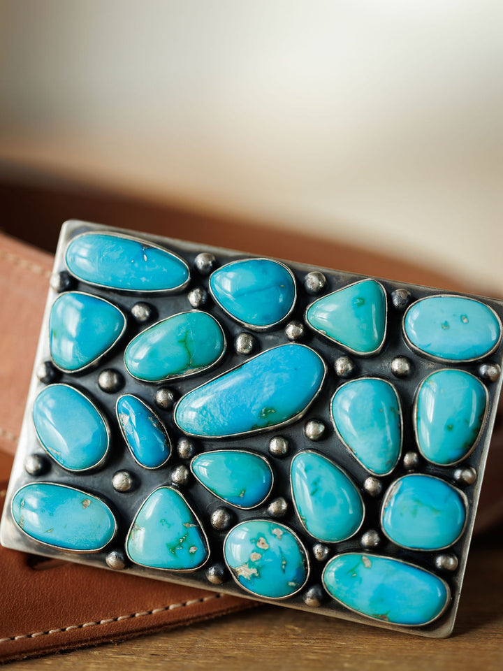 A close-up of the Amorita Buckle by Savannah Sevens western life{&}style, crafted in sterling silver and embellished with uniquely shaped turquoise stones, set against a blurred brown leather strap background.