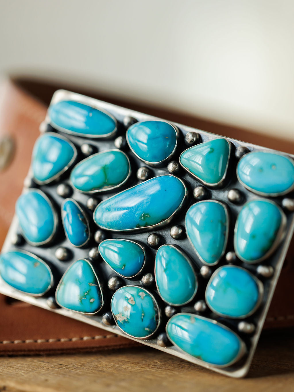 A close-up of the Amorita Buckle by Savannah Sevens western life{&}style, featuring variously shaped Sonoran turquoise stones set in sterling silver on a brown leather belt.