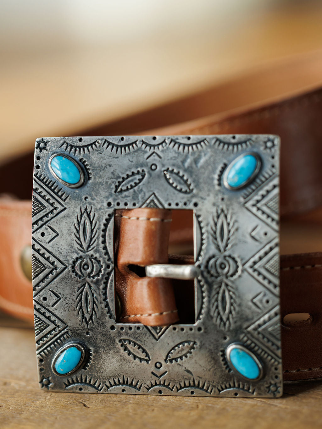 A close-up shows the Coalinga Buckle by Savannah Sevens western life{&}style: a brown leather belt with a square sterling silver buckle, engraved patterns, and four turquoise stones. The background is softly blurred.