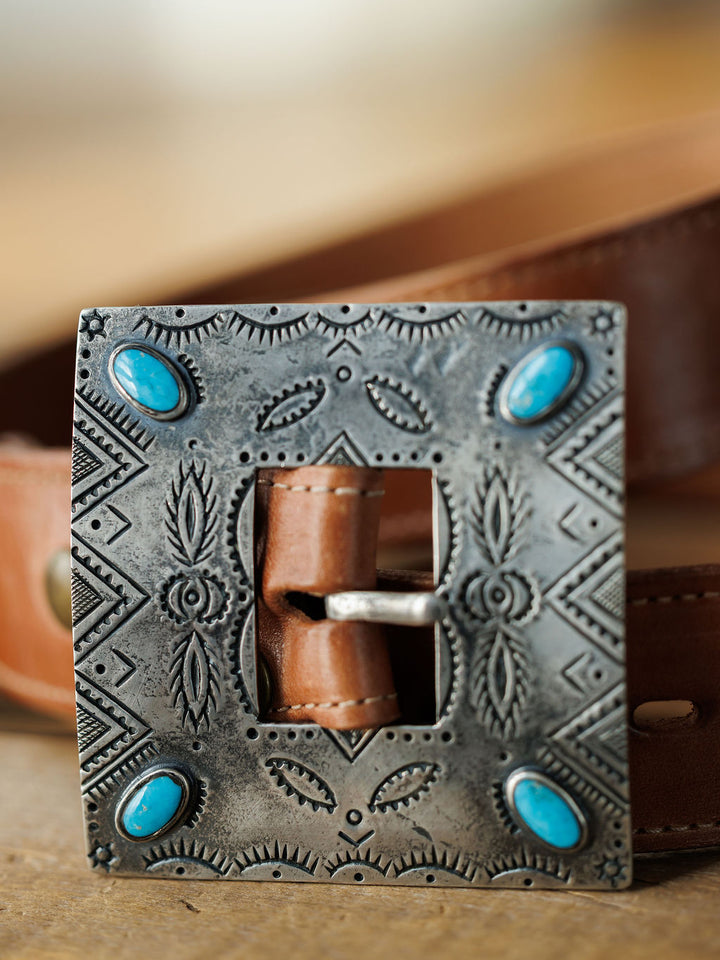 A close-up shows the Coalinga Buckle by Savannah Sevens western life{&}style: a brown leather belt with a square sterling silver buckle, engraved patterns, and four turquoise stones. The background is softly blurred.