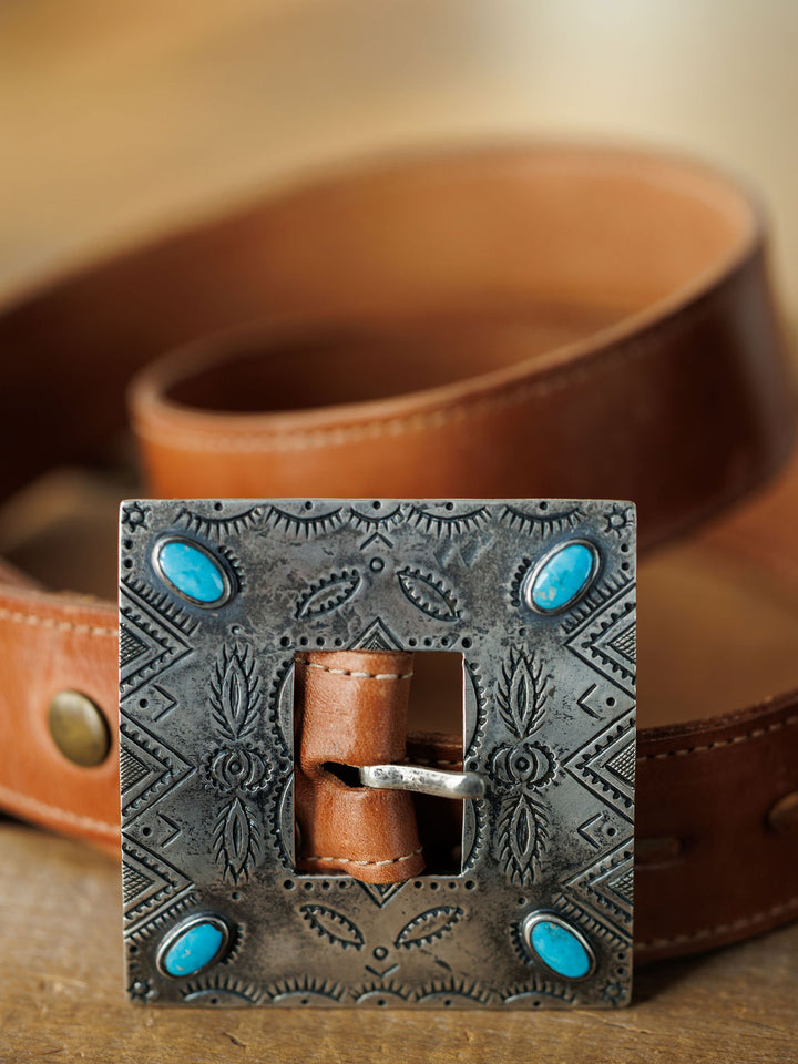 A close-up of the Coalinga Buckle by Savannah Sevens western life{&}style: a brown leather belt with a large handmade sterling silver buckle, engraved patterns, and four turquoise stones, shown on a wooden surface.