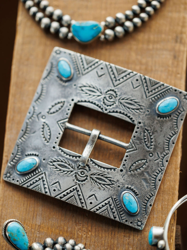 A close-up of the Coalinga Buckle by Savannah Sevens western life{&}style, a handmade sterling silver buckle with intricate engravings and five turquoise stones, displayed with beaded silver and turquoise jewelry on wood.