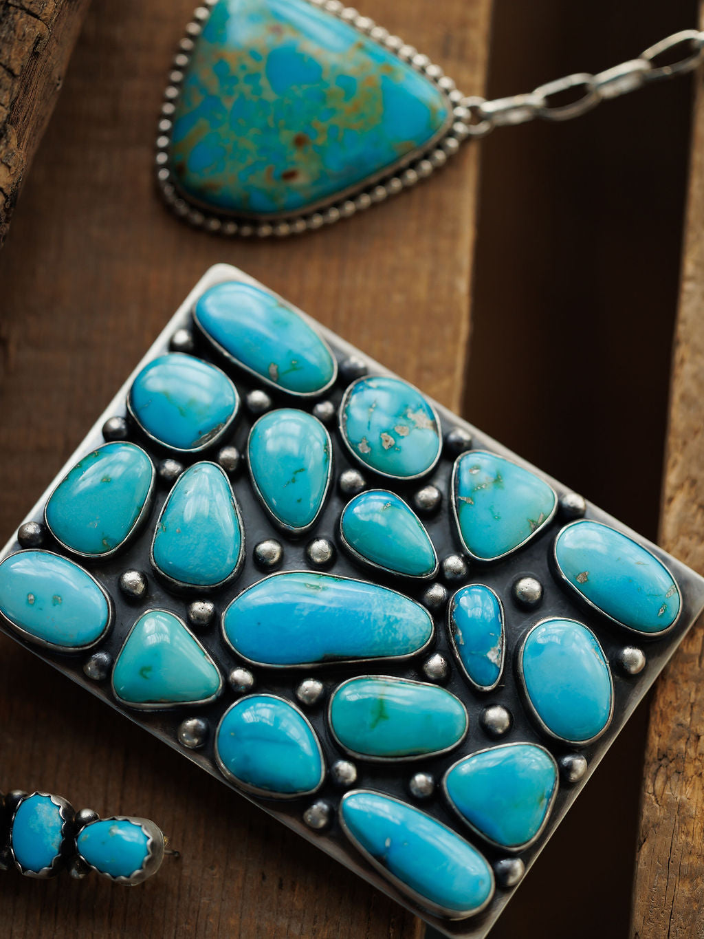The Amorita Buckle by Savannah Sevens western life{&}style, paired with a matching pendant necklace and earrings featuring polished turquoise, is displayed on a rustic wooden surface.