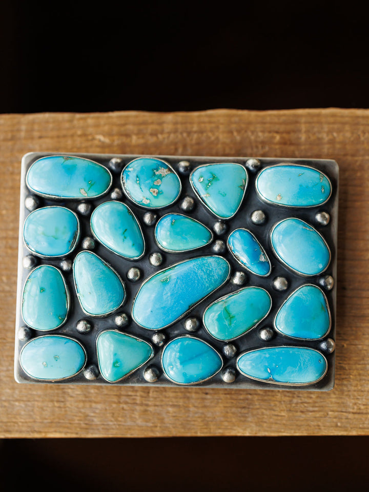 The Amorita Buckle by Savannah Sevens western life{&}style features a rectangular sterling silver design with irregular Sonoran turquoise stones and silver beads, displayed on a wooden surface.