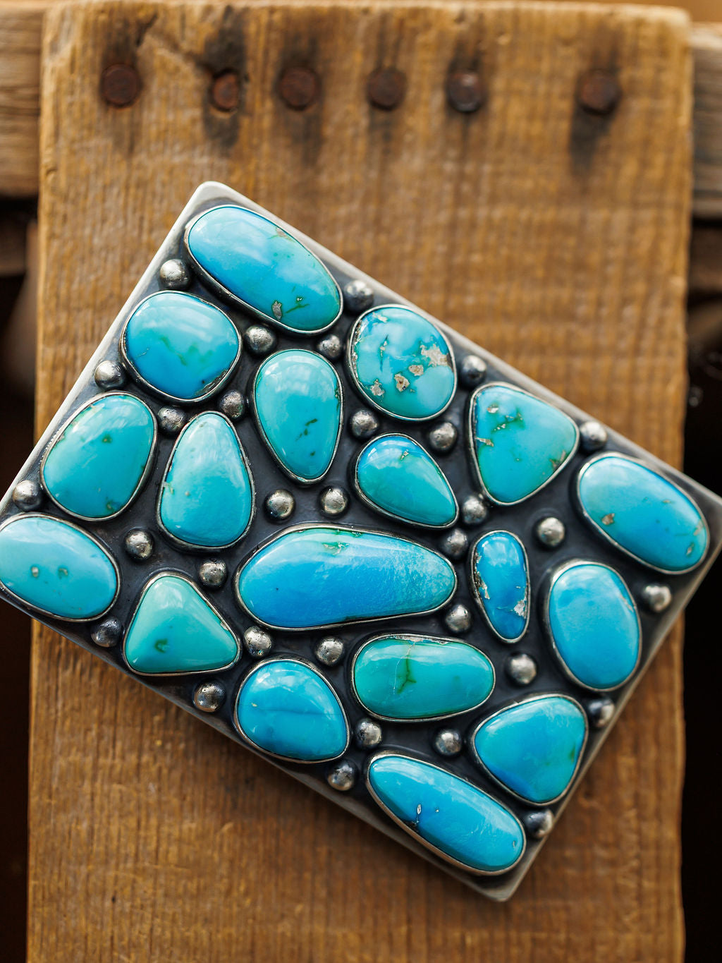 The Amorita Buckle by Savannah Sevens western life{&}style is a rectangular sterling silver buckle adorned with polished turquoise stones in various shapes and sizes, displayed on a rustic wooden background.