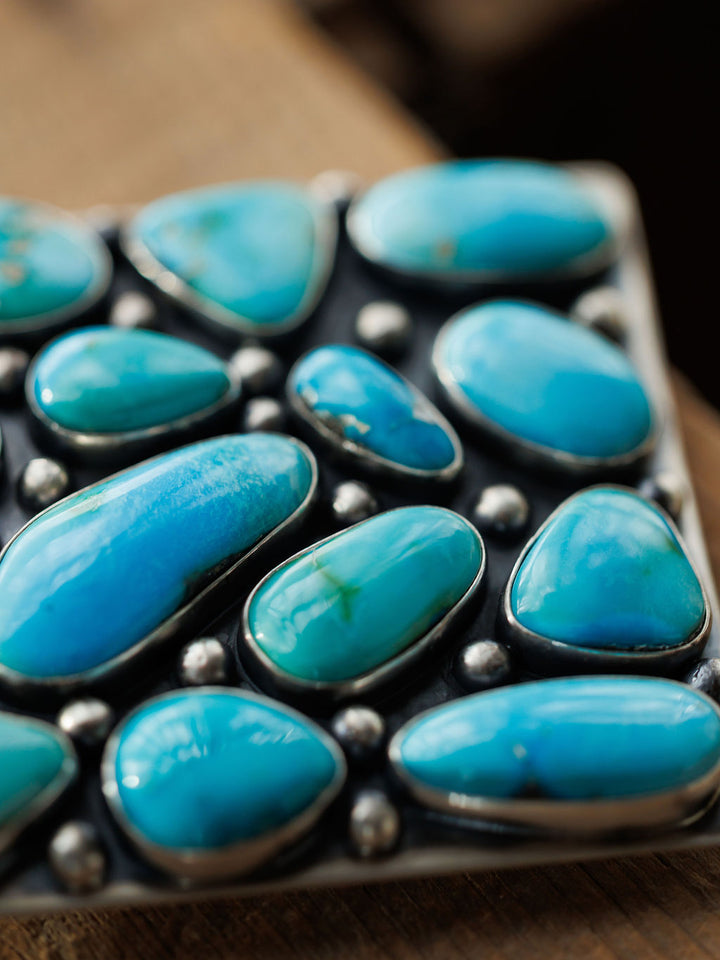 A close-up of polished turquoise stones from the Amorita Buckle by Savannah Sevens western life{&}style, set in sterling silver. The stones display smooth surfaces and vibrant blue-green hues against a dark background.