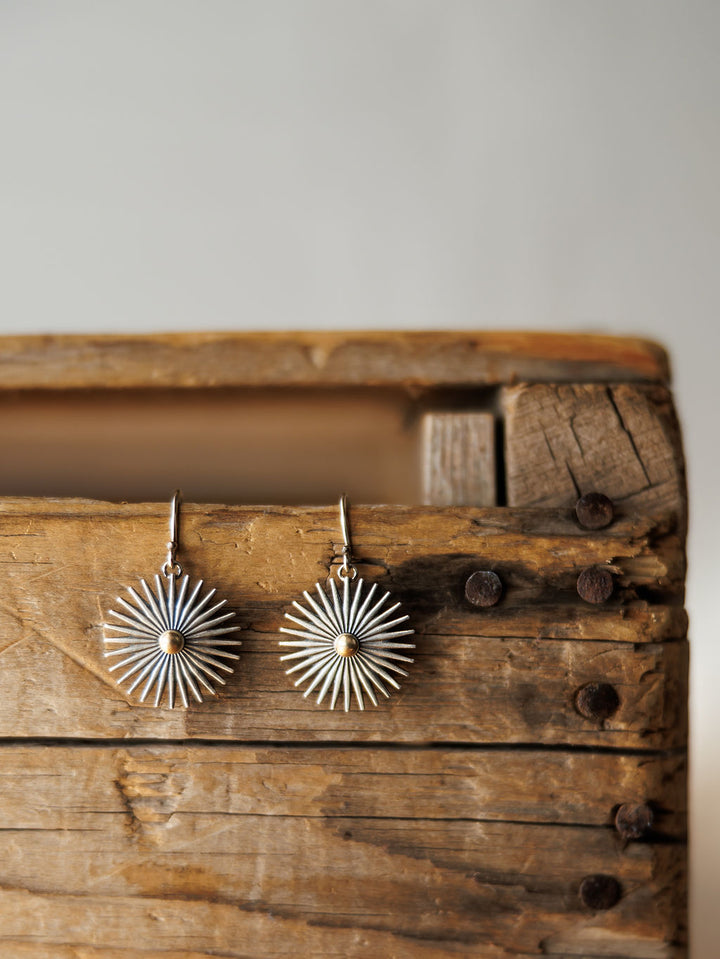 A pair of Rowel Earrings {Small} by Savannah Sevens western life{&}style, featuring sterling silver radiating spokes, rest on a weathered wooden box with visible nails and cracks; a softly blurred background highlights their handmade beauty.