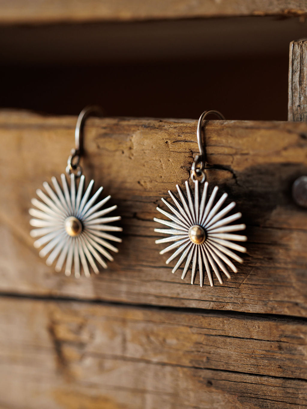 The Rowel Earrings {Small} by Savannah Sevens western life{&}style feature round, sunburst-shaped sterling silver with radiating spokes and a gold center, shown hanging from a rustic wooden surface.