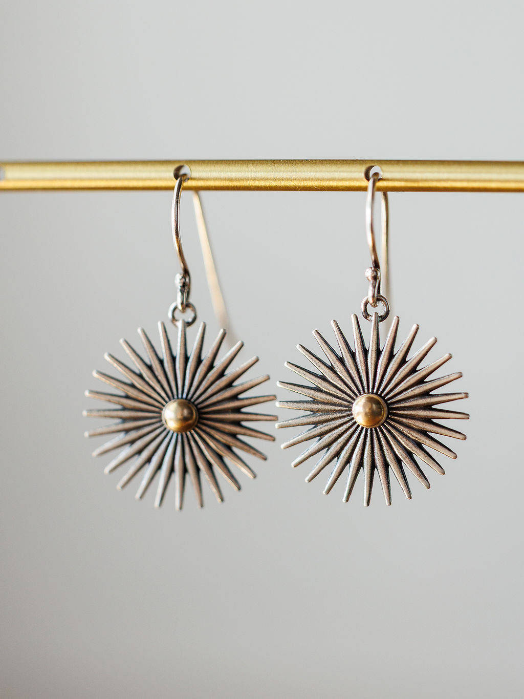 A pair of Rowel Earrings {Small} by Savannah Sevens western life{&}style, sterling silver with a sunburst design, radiating spikes, and a gold-colored center, hang from a gold bar against a neutral background.
