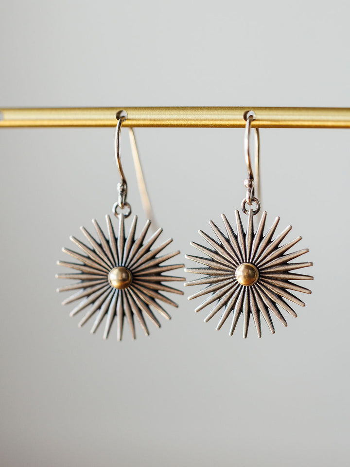 A pair of Rowel Earrings {Small} by Savannah Sevens western life{&}style, sterling silver with a sunburst design, radiating spikes, and a gold-colored center, hang from a gold bar against a neutral background.