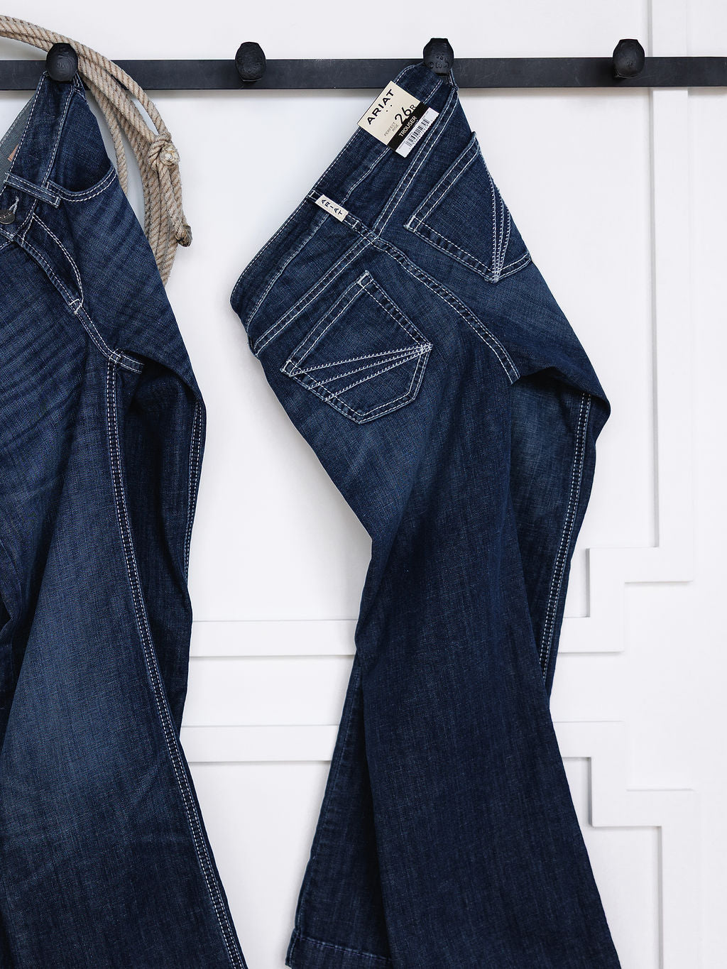 Two pairs of The Tripp Perfect Rise Trouser Jeans by Savannah Sevens western life{&}style hang on black hooks against a white paneled wall, with a beige rope visible on the left. They feature a comfy no-gap waist for extra comfort.