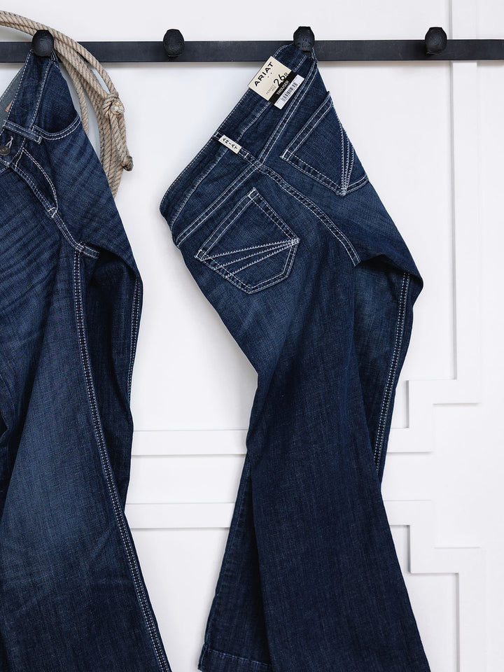 Two pairs of The Tripp Perfect Rise Trouser Jeans by Savannah Sevens western life{&}style hang on black hooks against a white paneled wall, with a beige rope visible on the left. They feature a comfy no-gap waist for extra comfort.