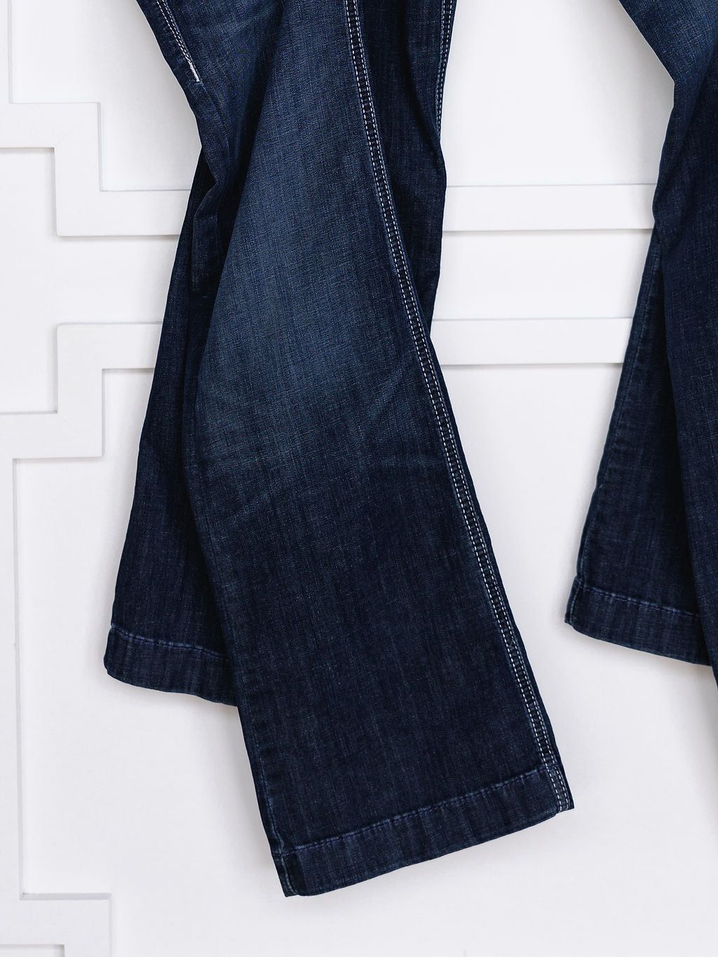 A close-up of the lower legs of The Tripp Perfect Rise Trouser Jean by Savannah Sevens western life{&}style, featuring a comfy no-gap waist and medium dark wash denim, shown hanging against a white wall with geometric patterns.