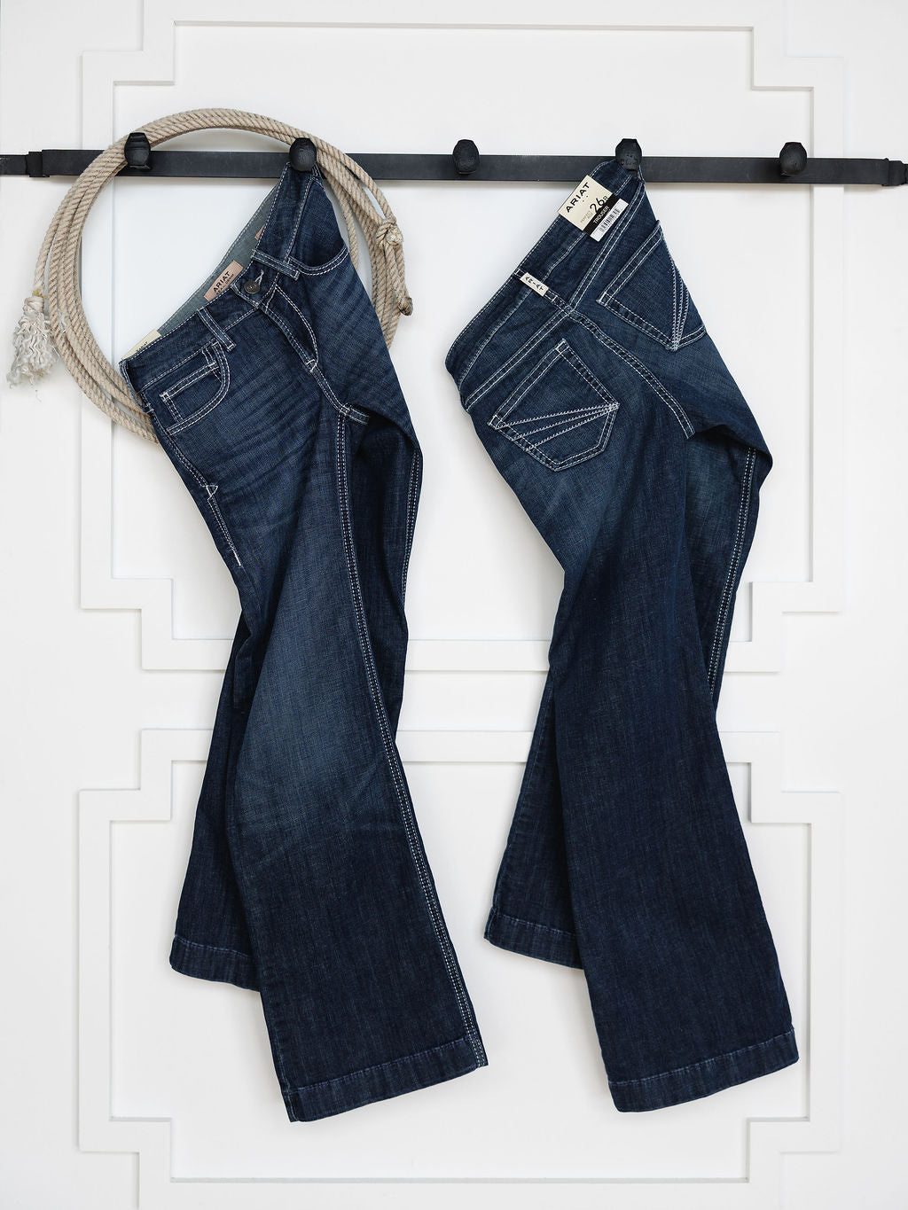 Two pairs of The Tripp Perfect Rise Trouser Jean by Savannah Sevens western life{&}style in a medium dark wash hang on a black wall rack, with a coiled rope on the left and a white wall with decorative molding in the background.