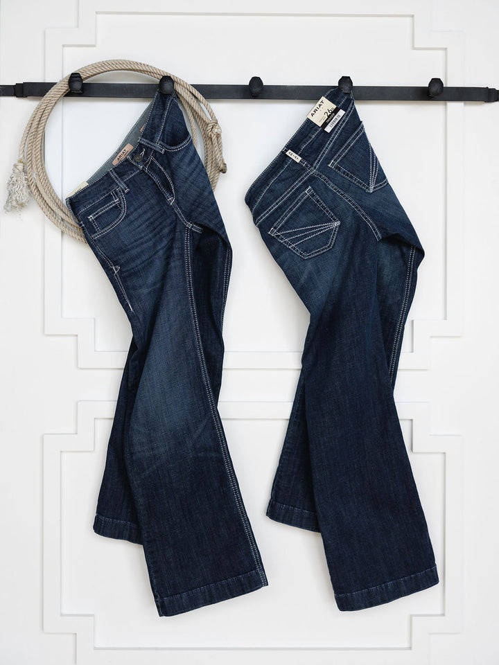 Two pairs of The Tripp Perfect Rise Trouser Jean by Savannah Sevens western life{&}style in a medium dark wash hang on a black wall rack, with a coiled rope on the left and a white wall with decorative molding in the background.
