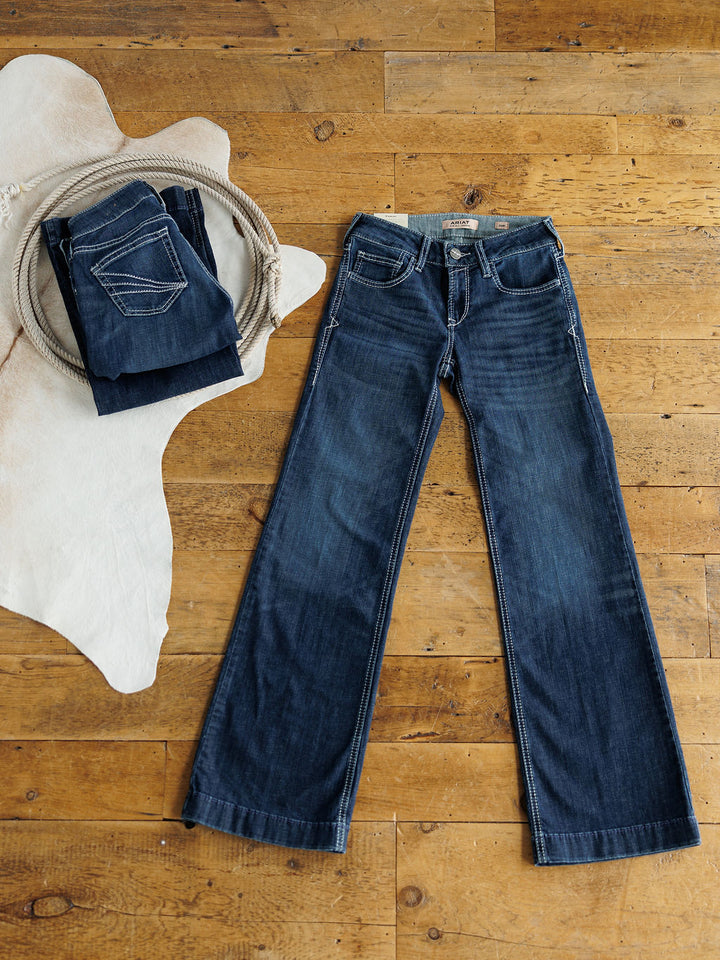 A pair of Savannah Sevens western life{&}style The Tripp Perfect Rise Trouser Jeans in a medium dark wash with a comfy no-gap waist, laid flat on a wooden floor beside folded jeans on a light cowhide rug.