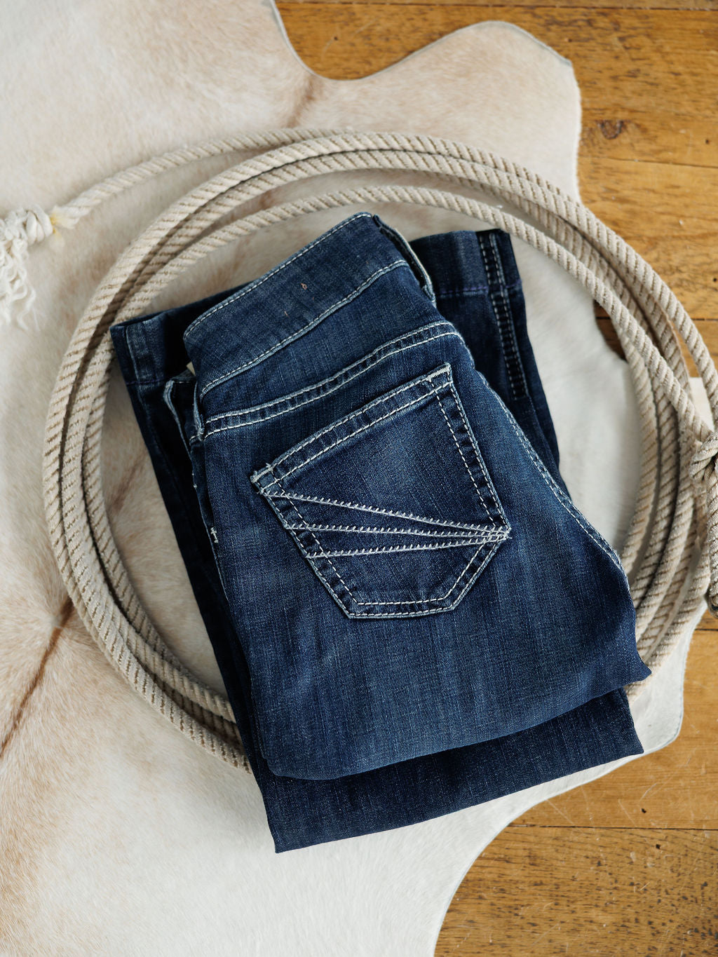 The Tripp Perfect Rise Trouser Jean by Savannah Sevens western life{&}style, in a medium dark wash, is folded on a cowhide rug with a coiled rope nearby. These jeans feature a comfy no-gap waist for all-day wear.